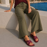 Marsala | Model wearing green pants and red wine-colored leather flat sandals against a coastal beach background