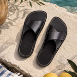 Nero | Black leather flat sandals against a beach background