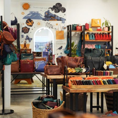 A retail display of leather handbags, backpacks, and wallets arranged on tables and hanging racks inside a boutique. The scene includes shelves with colorful accessories, a wall mural with travel-themed illustrations, and baskets of additional goods.