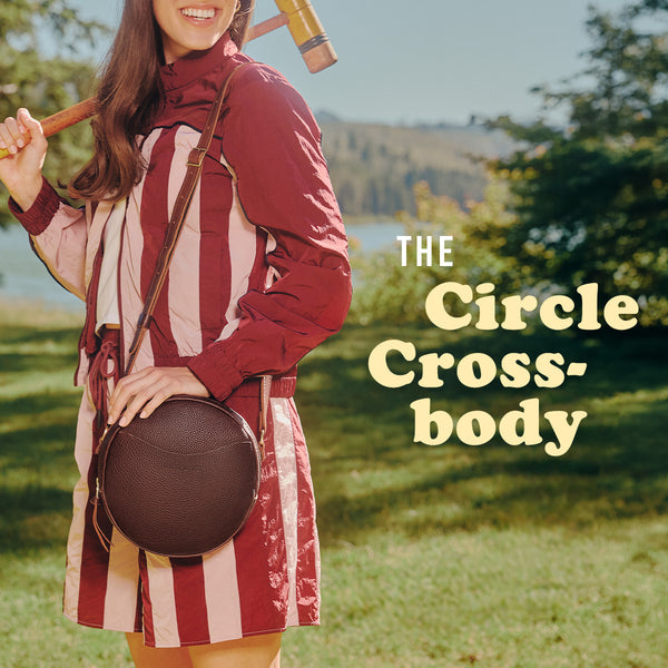 A woman outdoors in a grassy park holding a cricket ball mallet and carrying a round brown crossbody bag, wearing a burgundy and cream vertically striped dress. Text on the right reads 'The Circle Crossbody'.