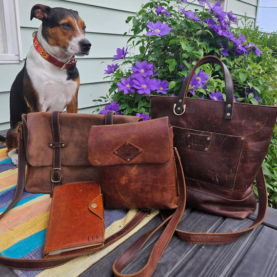Dog sitting behind archive leather bags and a wallet on a wooden surface with flowers in the background