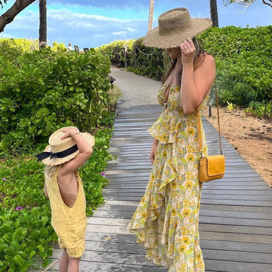 An adult and a child in yellow outfits walking on a wooden path with greenery around wearing a Metro Crossbody in Sunflower.