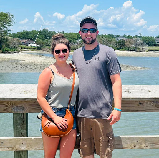 Two people standing on a wooden pier by a body of water with trees and houses in the background.