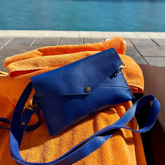 Envelope Convertible Crossbody in blue laying on an orange towel by the pool