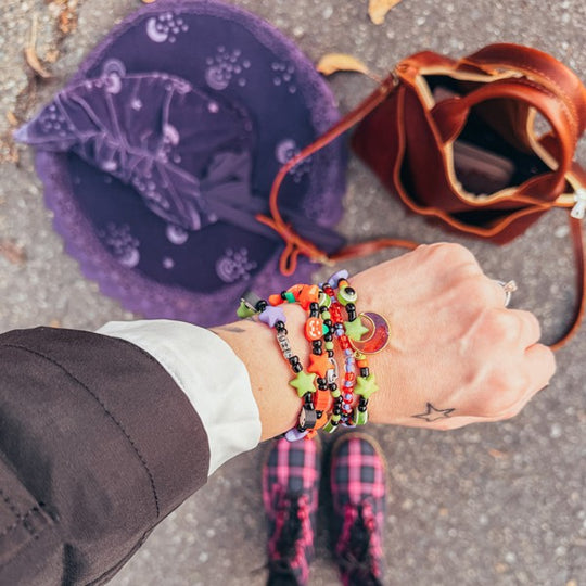 A wrist full of bracelets with a witch hat and leather bag in the background