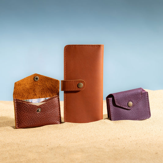 Three leather wallets in brown and purple on a sandy surface with a blue sky background.