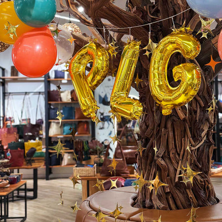 Gold foil balloon letters spelling 'PLG' hanging on a decorative tree trunk in a party setting, with colorful balloons and gold star streamers in a shop interior