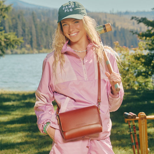A woman outdoors by a lake wearing a pink tracksuit and green cap with 'CAMP PLG' text, wearing a Madrone Metro Crossbody bag.