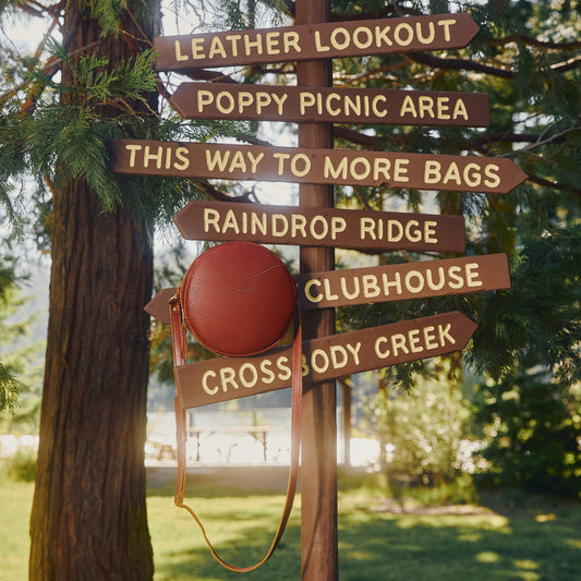 A brown leather circular shoulder bag with a tan strap hangs from a rustic wooden signpost outdoors. The signpost points to destinations including Leather Lookout, Poppy Picnic Area, This Way to More Bags, Raindrop Ridge, Clubhouse, and Cross Body Creek, with a tree and greenery in the background.