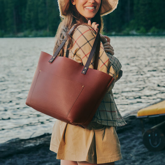 A woman outdoors by the water wearing a wide-brimmed hat and carrying a large brown leather tote bag over her shoulder.