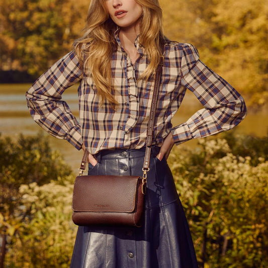 A woman outdoors wearing a brown leather crossbody bag with a matching strap, styled with a beige and navy plaid shirt and a dark skirt.