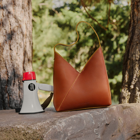 A brown pebbled leather shoulder bag with a single strap, styled on a stone ledge outdoors next to a white and red megaphone, with trees and green foliage in the background.