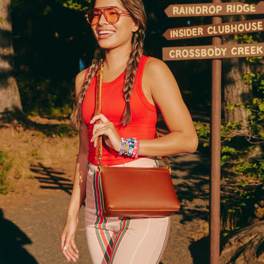 A woman in a red tank top and striped pants carrying a brown leather crossbody bag, standing outdoors next to a wooden signpost with directions to Raindrop Ridge, Insider Clubhouse, and Crossbody Creek.