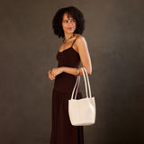 Bone Classic | Model wearing sleeveless dark brown dress carrying petite bucket shaped tote bag with matching leather handles against dark grey background