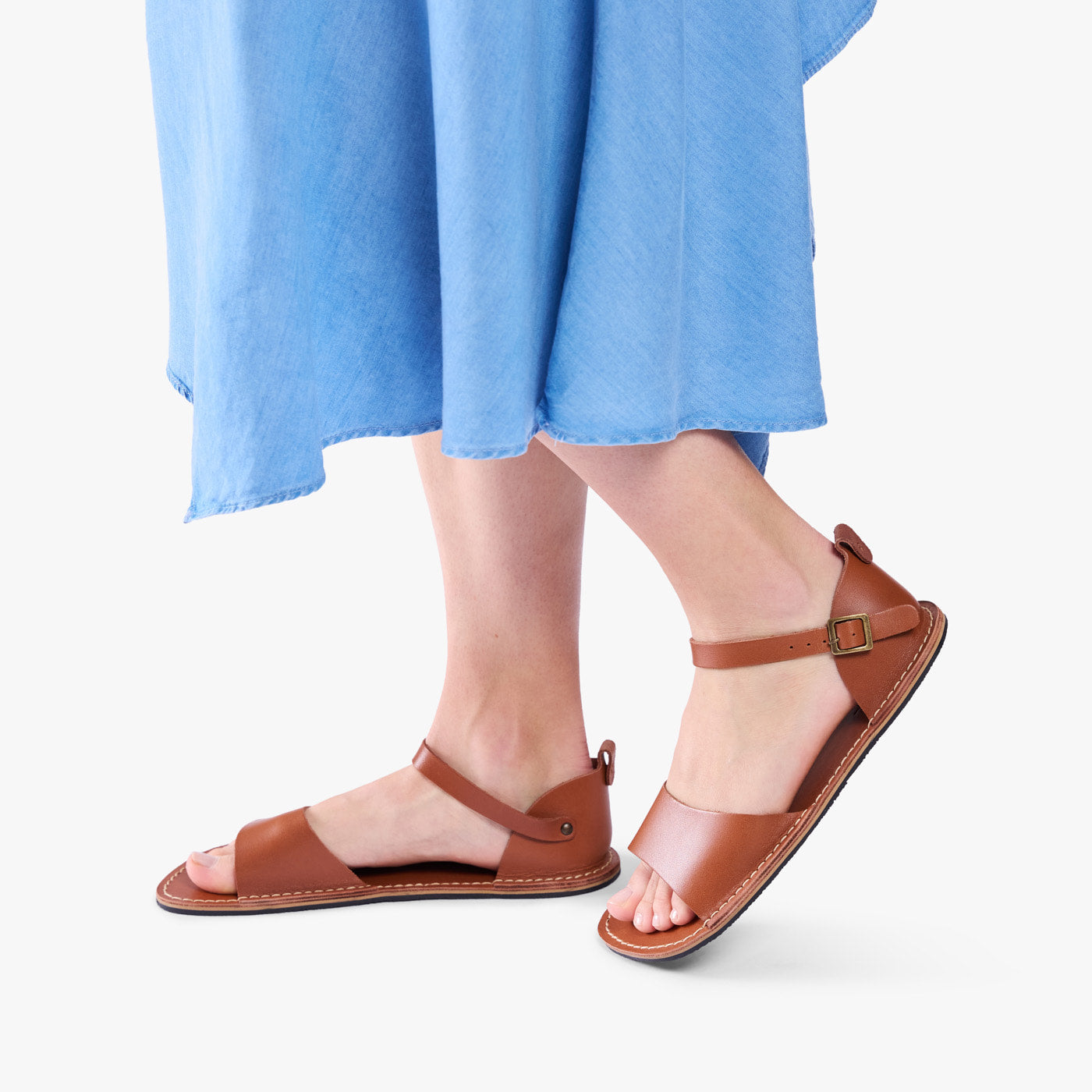 Miele | Person wearing blue dress and tan flat sandals with an ankle strap and buckle, shown against a plain background.