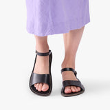 Nero | Person wearing purple dress and black flat sandals with an ankle strap and buckle, shown against a plain background.