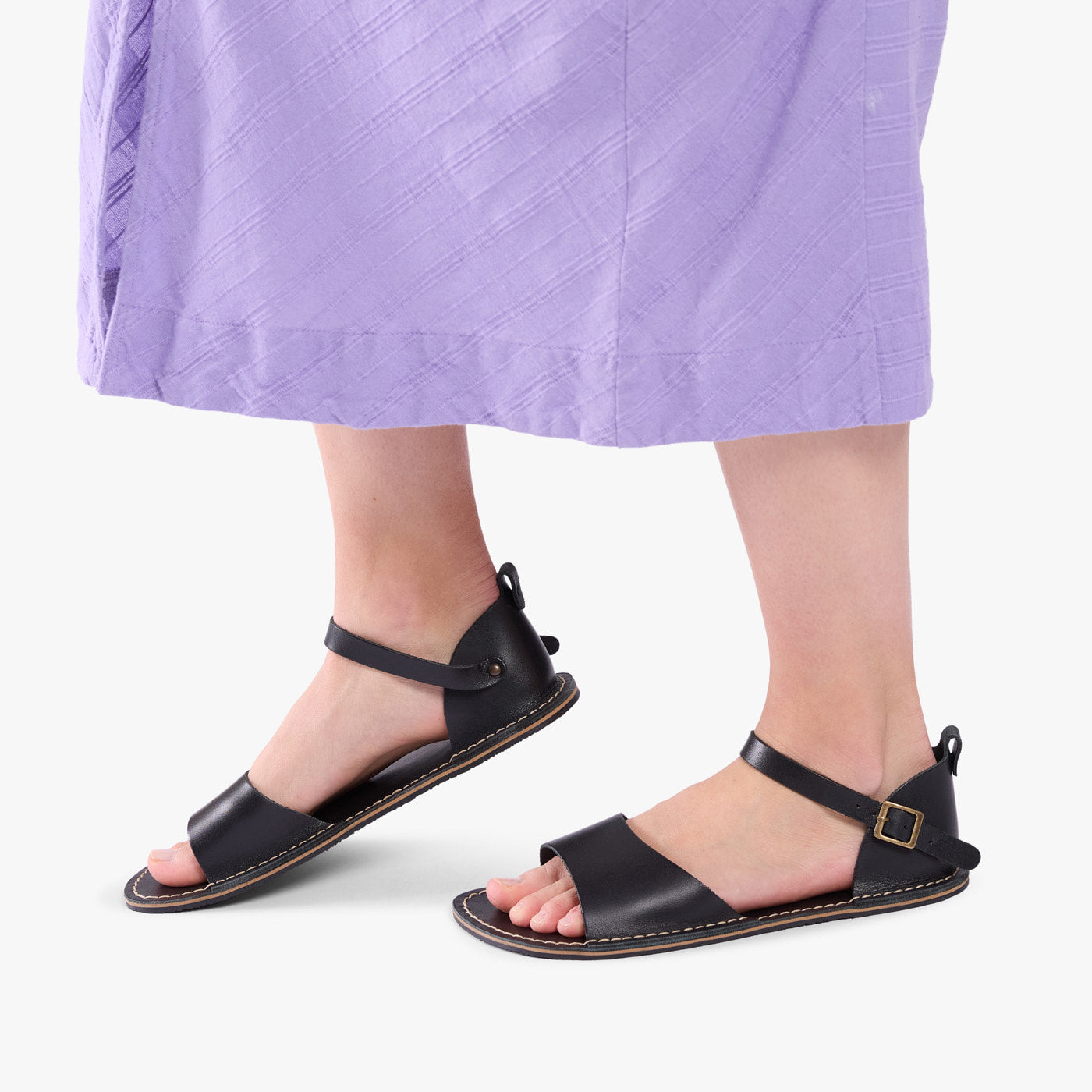 Nero | Person wearing purple dress and black flat sandals with an ankle strap and buckle, shown against a plain background.