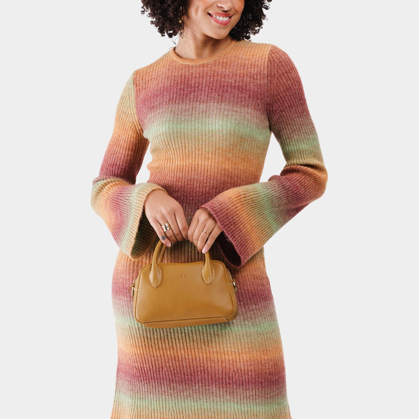 Mango*Small | A bowler styled bag with a crossbody strap being held by a model wearing a multi-colored dress