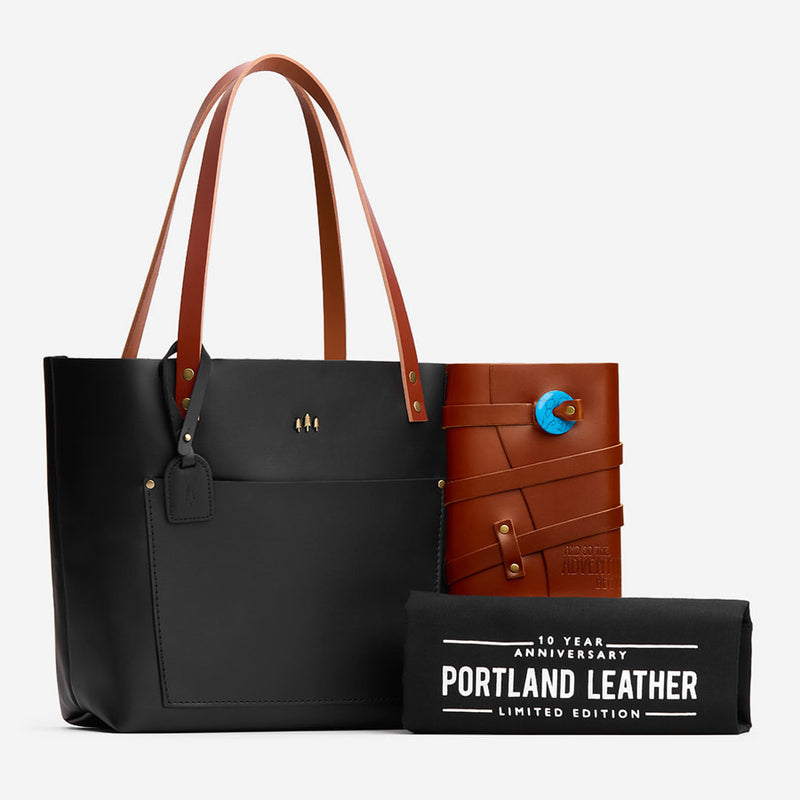 Black leather tote bag with brown handles and a Wrap Journal on a white background, featuring 'Portland Leather' branded dust cover.