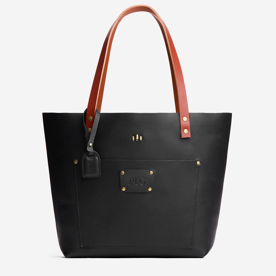 Black tote bag with brown handles and custom monogram plate on a light gray background