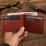 Marrone | A hand holds an open leather bifold wallet showing card slots and cash on a wooden tabletop in the background