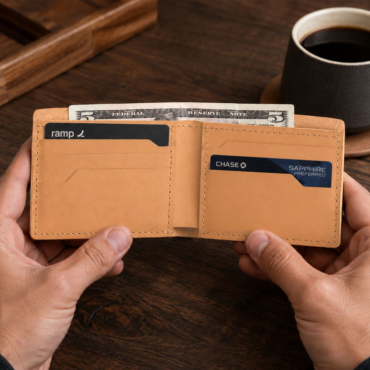 Naturale | A leather bifold wallet held open by two hands over a dark wooden table, with cash and cards visible inside. 
