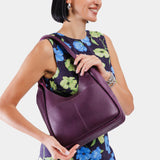 Plum Large | A model holding a u-shaped, flat-bottomed leather shoulder bag with two outside pockets