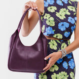 Plum Large | A model holding a u-shaped, flat-bottomed leather shoulder bag with two outside pockets