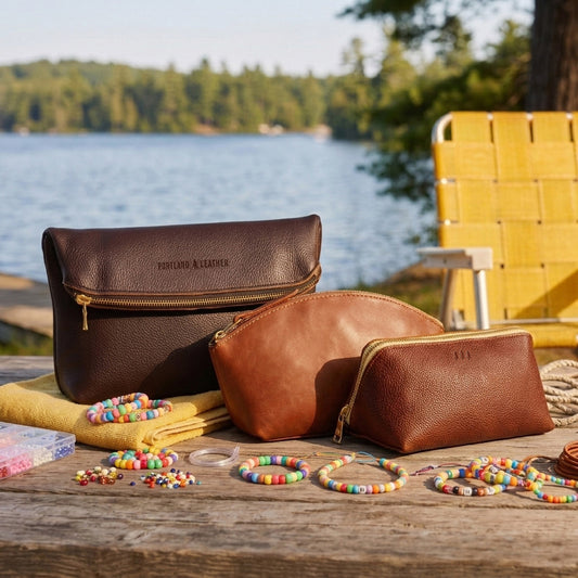 Three brown leather zip pouches of different sizes arranged on a wooden table outdoors, with colorful beaded bracelets and folded yellow towels in front. A lake and trees are visible in the background, along with a yellow chair.