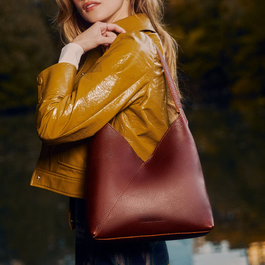 A woman wearing a glossy mustard yellow jacket carries a Nutmeg Large Naomi Shoulder Bag. The background is a soft, blurred outdoor scene with green foliage.
