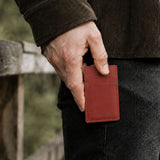 Marrone | A close-up of a model's hand holding a small red leather card holder against their dark jeans, with a blurred outdoor background.
