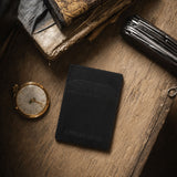 Nero | A black leather card holder styled on a rustic wooden table with vintage books, a pocket watch, and a folding knife.