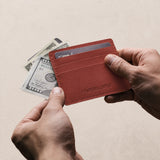 Marrone | A card holder held by two hands over a light wood table, with a credit card and money