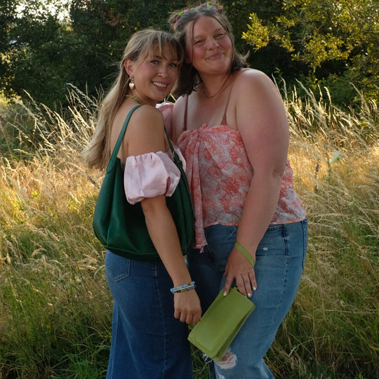 Two women posing in front of the woods with tall grass carrying a green leather shoulder bag and a light green leather wristlet wallet.