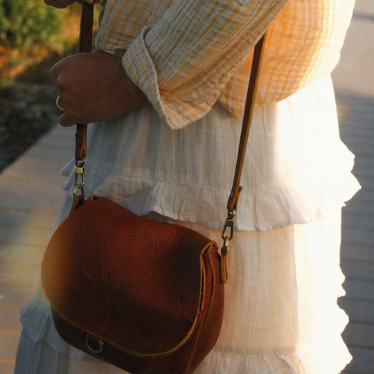 Woman wearing a white skirt and yellow striped top carrying a brown leather crossbody bag with straps and a front buckle