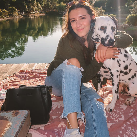 Women sitting on a picnic blanket hugging a dalmatian dog next to a lake and a black leather bucket bag
