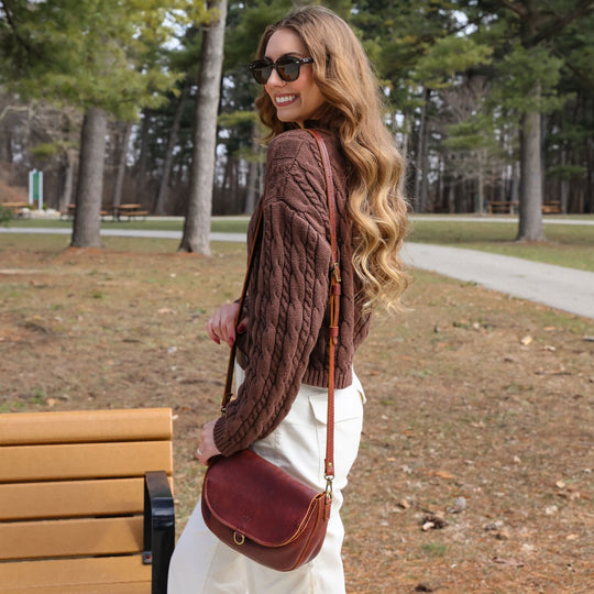 Woman wearing sunglasses and a brown knitted sweater carrying a brown leather crossbody bag with a front buckle in front of a bench in a forested park