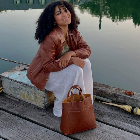 Woman wearing a leather jacket and a white skirt with a brown leather tote bag sitting in front of a lake background