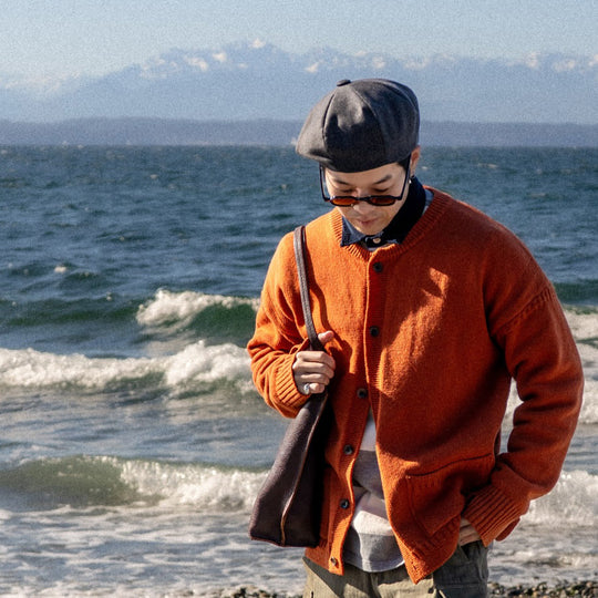 Man wearing a beret, tinted glasses, and an orange button up cardigan carrying a leather shoulder bag with ocean waves in the background.