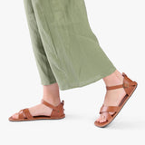 Miele | Close-up of model wearing light green pants and brown flat sandals with an ankle strap and buckle, shown against a plain background