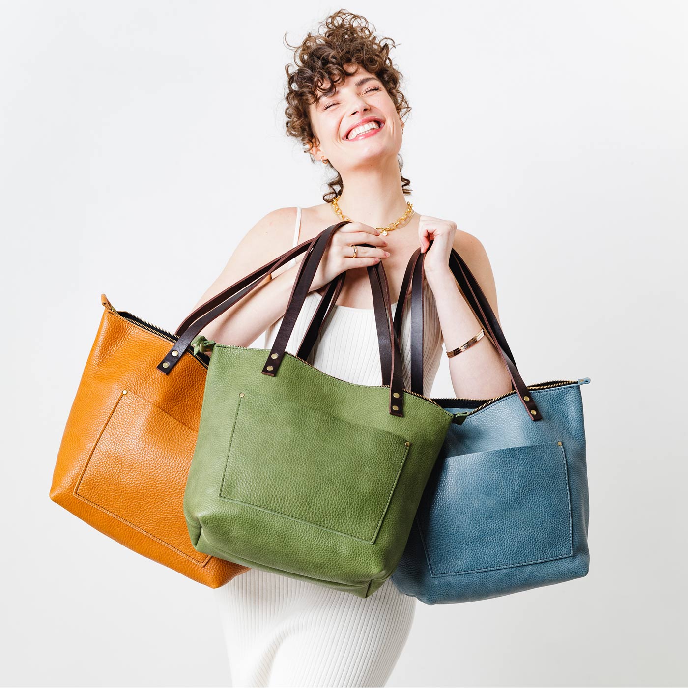 1 Mystery Tote | Model holdingan orange, light green, and light blue leather tote bags with front pocket. 